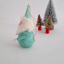 Load image into Gallery viewer, Another side view of a mint green, spun cotton gnome, standing on a white surface with small Christmas trees in the background.