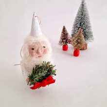 Load image into Gallery viewer, A spun cotton gnome holding a wreath, standing on a white surface with small bottle brush trees in the background.
