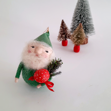 Load image into Gallery viewer, Decorative spun cotton gnome ornament with a green hat, holding a red mushroom, standing on a white background with bottle brush trees in the distance.