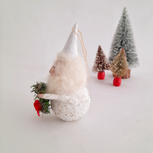 Load image into Gallery viewer, The side view of a spun cotton gnome holding a wreath, standing in front of small Christmas trees on a light background.