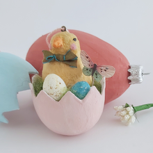 Load image into Gallery viewer, A close-up view of a spun cotton Easter chick in a cracked egg. Blue and muted red egg ornaments sit behind it. Pic 2 of 7.