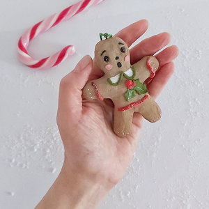 A hand holding a vintage style, spun cotton gingerbread girl ornament. A candy cane lays in the background. Pic 4 of 8. 