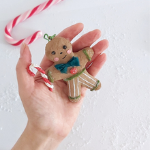 Load image into Gallery viewer, A hand holding a vintage style spun cotton gingerbread man ornament against a white background. A candy cane is laying on the table. Pic 2 of 8.