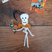 Load image into Gallery viewer, A vintage style, spun cotton pipe cleaner skeleton laying on a dark wood background next to a vintage Halloween illustration. Pic 1 of 5.