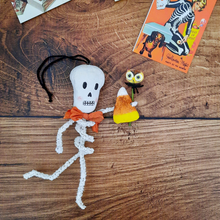 Load image into Gallery viewer, A vintage style, spun cotton pipe cleaner skeleton laying on a wooden background next to a vintage Halloween advertisement. Pic 1 of 5.