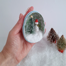 Load image into Gallery viewer, A hand holding a vintage style, spun cotton snowman diorama ornament against a white background. Two vintage bottlebrush trees sit nearby. Pic 2 of 6.