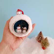 Load image into Gallery viewer, A hand holding a diorama ornament of vintage style snowmen against a light blue background, above two vintage bottle brush trees. Pic 3 of 5.