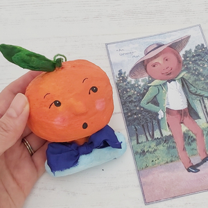 Spun cotton orange boy held in hand, next to Victorian illustration of anthropomorphic orange. Pic 2 of 6.