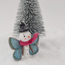 Cargar imagen en el visor de la galería, A side view of spun cotton snowman butterfly ornament, standing next to mini Christmas tree. Pic 3 of 5.