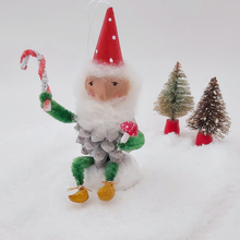 Load image into Gallery viewer, Vintage style spun cotton pine cone elf holding a spun cotton mushroom and pipe cleaner candy cane. He sits on a mound of spun cotton snow against a white background with two vintage bottle brush trees in the distance. Pic 2 of 9.