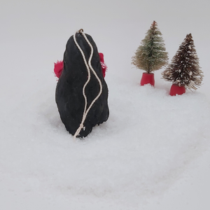 Back view of spun cotton pine cone penguin sitting on fake snow with two bottle brush trees in the distance. Pic 9 of 10. 