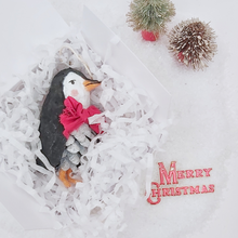 Load image into Gallery viewer, Vintage style spun cotton pine cone penguin laying in white gift box amongst white tissue shredding. Two miniature bottle brush trees and a vintage Merry Christmas decoration surround the box. Pic 7 of 10.