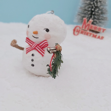 Load image into Gallery viewer, Close up of garland and silver jingle bells that the spun cotton snowman is holding. He's sitting on fake snow against a light blue background with a bottle brush tree and vintage Merry Christmas decoration in the distance. Pic 4 of 7.