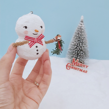 Load image into Gallery viewer, Vintage style spun cotton snowman ornament, held in hand for size comparison. He&#39;s against a light blue background with a bottle brush tree and vintage Merry Christmas sign in the distance. Pic 1 of 7.
