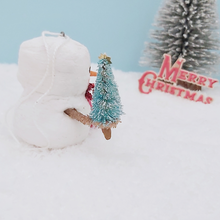 Load image into Gallery viewer, Side view of vintage style spun cotton snowman, sitting on snow with bottle brush tree and Merry Christmas sign  in distance. Pic 7 of 8.