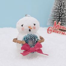 Load image into Gallery viewer, Close up view of spun cotton snowman and his wreath. He's against a light blue background, sitting on snow, with a bottle brush tree in the distance. Pic 4 of 8.