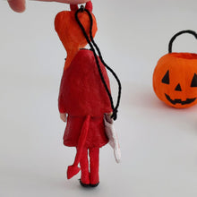 Load image into Gallery viewer, The back side of a spun cotton devil girl ornament is shown, including her devil tail. A blurred orange jack-o'-lantern bucket sits in the background on a plain light surface.

