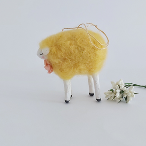 A side view of a yellow, vintage style needle felted spun cotton sheep ornament. White flowers sit behind her, against a white background. Pic 5 of 7. 