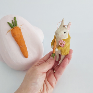 A vintage style spun cotton bunny ornament being held in hand against a white background. A light pink paper mache egg box sits in the distance. Pic 2 of 12. 