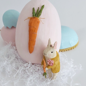 A vintage style, spun cotton bunny ornament leaning against a light pink paper mache egg adorned with a spun cotton carrot. Pastel eggs sit in the distance. Pic 12 of 12. 