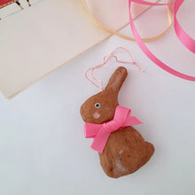 Load image into Gallery viewer, A brown spun cotton bunny, wearing a bright pink bow, lays on a white surface next to a vintage Easter card and colourful ribbon. 
