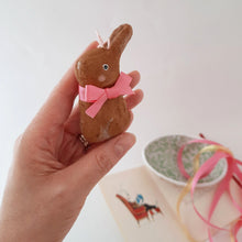 Load image into Gallery viewer, A hand holds a brown spun cotton bunny, that's wearing a pink bow. Pink and yellow ribbons and a paper mache egg sit in the light background. 
