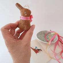 Load image into Gallery viewer, A hand holds a small brown spun cotton bunny, which wears a pink bow. Ribbons and a paper mache egg sit in the background.
