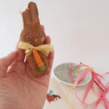 Load image into Gallery viewer, A hand holding a small spun cotton bunny,  that's wearing a yellow bow and holding a carrot. A vintage Easter card, paper mache egg and colourful ribbons lay in the background.
