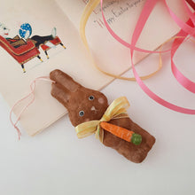 Load image into Gallery viewer, An image of a brown, spun cotton bunny ornament, holding a carrot, on a white surface. A vintage Easter card and yellow and pink ribbons are in the background.
