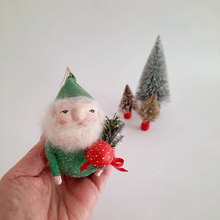 Load image into Gallery viewer, Hand holding a gnome ornament  wearing a green hat and holding a red mushroom, with miniature bottle brush trees in the background.
