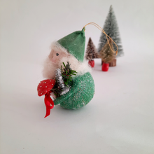 Load image into Gallery viewer, A side view of a spun cotton gnome resembling wearing a green hat and holding a red mushroom on a white background.
