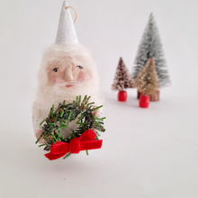 Load image into Gallery viewer, A closer view of a spun cotton gnome, holding a wreath with red bow, set against a light background with small Christmas trees.
