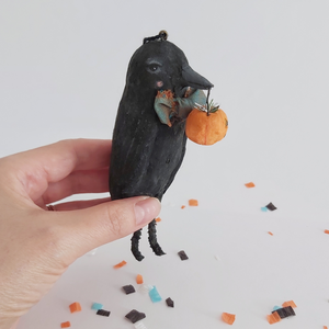 A vintage style spun cotton crow ornament, held in a hand over Halloween confetti against a white background. Pic 4 of 8. 