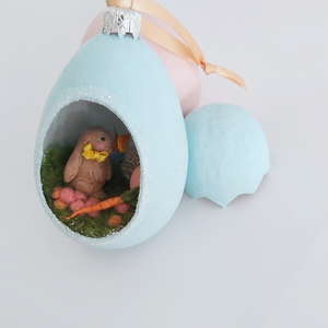 Another close-up of a different section of the vintage style spun cotton Easter bunny diorama ornament. It leans against pink and blue egg ornaments, on a white background. Pic 6 of 8. 