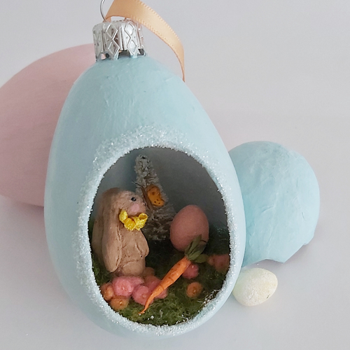 A vintage style spun cotton Easter bunny diorama ornament sitting next to pink and blue eggs against a white background. Pic 1 of 8. 