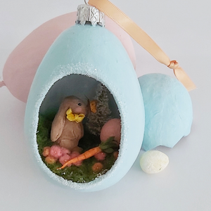 Another close-up of vintage style spun cotton Easter bunny diorama ornament. It lays against pink and light blue egg ornaments, on a white background. Pic 4 of 8. 