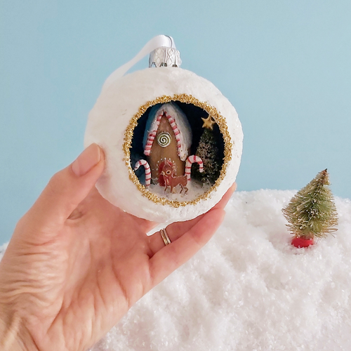 A hand holding a spun cotton gingerbread house diorama ornament against a light blue background over fake snow. A mini bottle brush tree sits in the distance. Pic 1 of 6. 