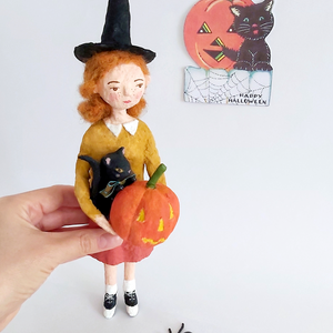 A hand holding a vintage style, spun cotton Halloween girl art doll. A vintage style Halloween greeting hangs in the background. Pic 2 of 7.