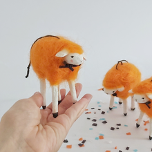 Load image into Gallery viewer, A vintage style, spun cotton needle felted orange sheep ornament held in a hand. Two orange sheep stand in the distance, on Halloween confetti, against a white background. Pic 2 of 5. 
