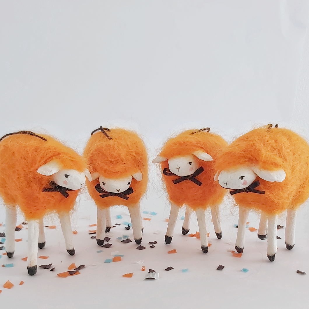 A close view of four vintage style, spun cotton needle felted orange sheep ornaments. They stand on Halloween confetti, against a white background. Pic 1 of 5. 