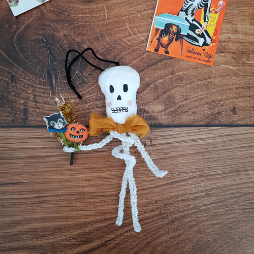 A vintage style, spun cotton pipe cleaner skeleton laying on a dark wood background next to a vintage Halloween illustration. Pic 1 of 5.