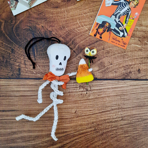 A vintage style, spun cotton pipe cleaner skeleton laying on a wooden background next to a vintage Halloween advertisement. Pic 1 of 5. 