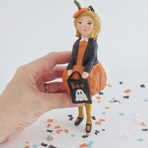 A vintage style spun cotton pumpkin girl ornament, held in hand on Halloween confetti against a white background. Pic 1 of 9