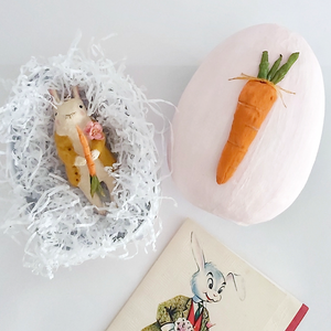 A vintage style, spun cotton bunny ornament laying in a light pink paper mache egg box with white tissue shredding. A vintage Easter card lays below in the photo. Pic 11 of 12. 
