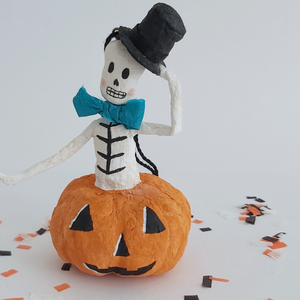 Another closer view of a vintage style, spun cotton skeleton in a jack-o-lantern. It's sitting on Halloween confetti against a white background. Pic 5 of 7. 