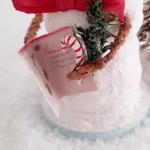 A close-up of a card, greenery, and spun cotton candy cane held by a vintage style snowman. Pic 1 of 6. 