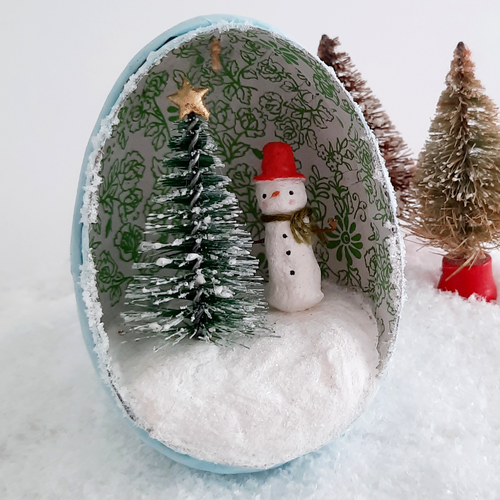 A close view of a vintage style, spun cotton snowman diorama ornament. Two vintage bottlebrush trees sit behind it. Pic 1 of 6. 