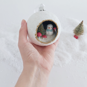 A vintage style spun cotton snowman diorama ornament held in hand over fake snow. A mini bottle brush tree sits in the distance. Pic 3 of 6.  