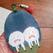 Cargar imagen en el visor de la galería, A wood ornament with two sheep on a blue background, held by a hand on a wooden surface. 
