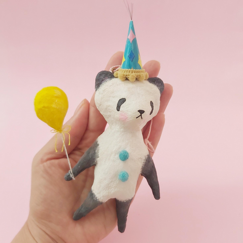 Spun Cotton Panda ornament, holding yellow balloon and wearing a colourful party hat. Held in a hand. Picture 1 